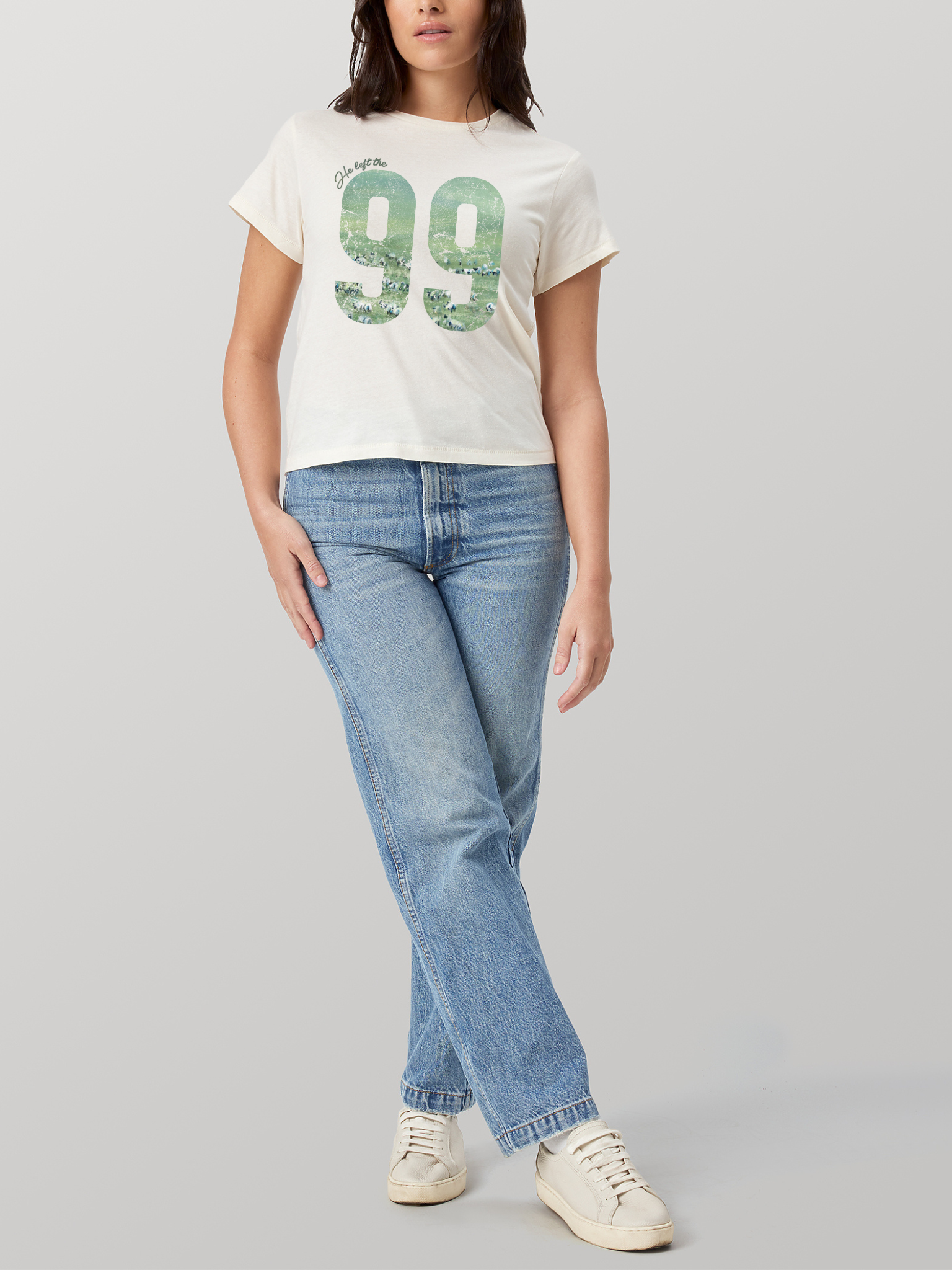 Woman wearing SĒK Apparel He Left the 99 Baby Tee in beige — Christian graphic tee with '99' sheep design, styled with jeans and white sneakers