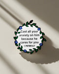 SĒK Apparel 1 Peter 5:7 enamel pin — floral Christian lapel pin with 'Cast all your anxiety on Him' scripture, front view on white background