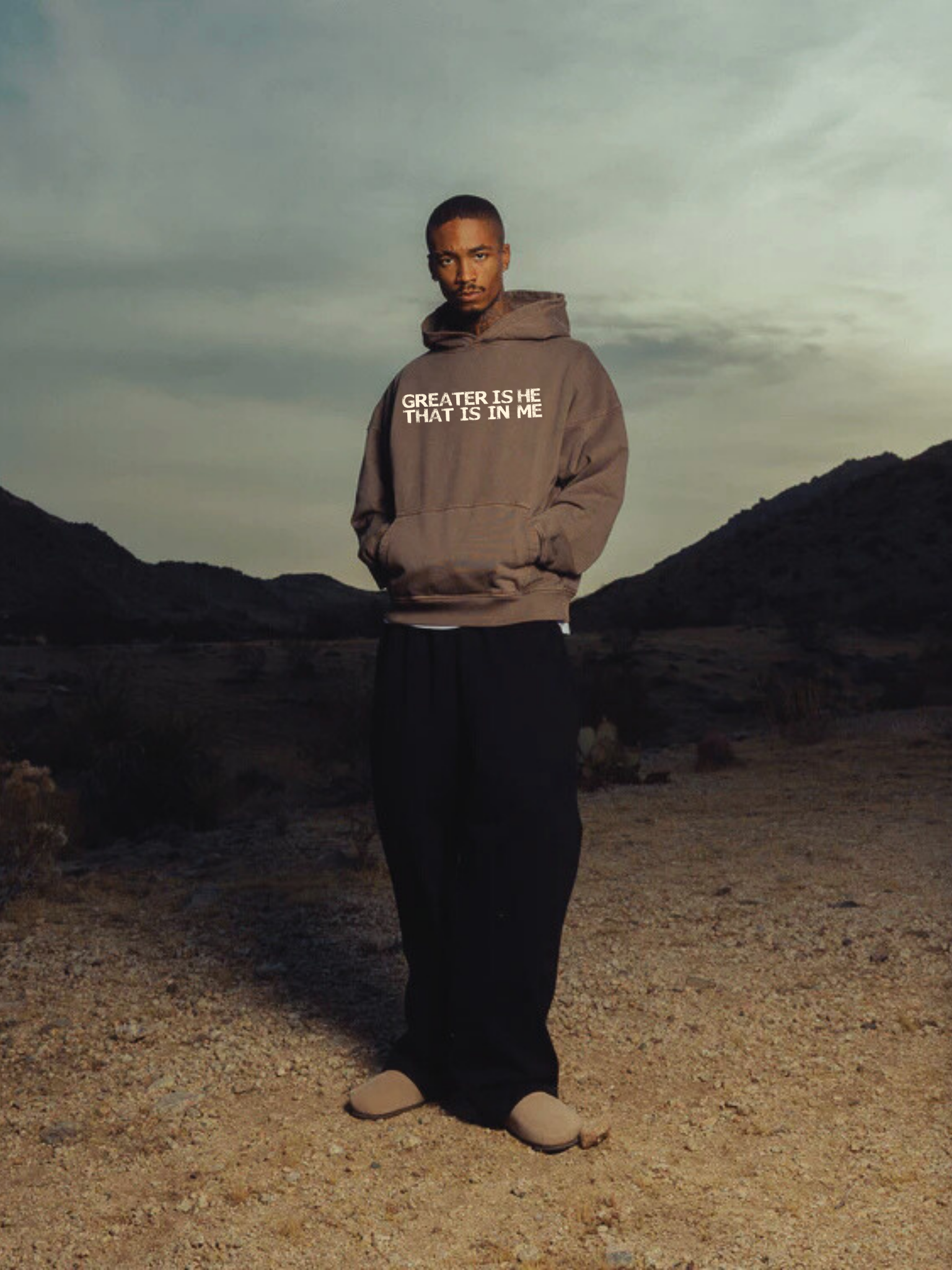 Male model wearing SĒK Apparel Greater is He Hoodie in brown — front view