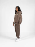 Woman wearing SĒK Apparel Greater is He Hoodie in brown — front view