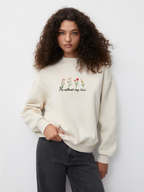 Woman wearing the SĒK Apparel He Restores Crewneck in beige — front view of faith-inspired Christian sweatshirt with 'He Restores' embroidered text and floral detail, referencing Psalm 23:3