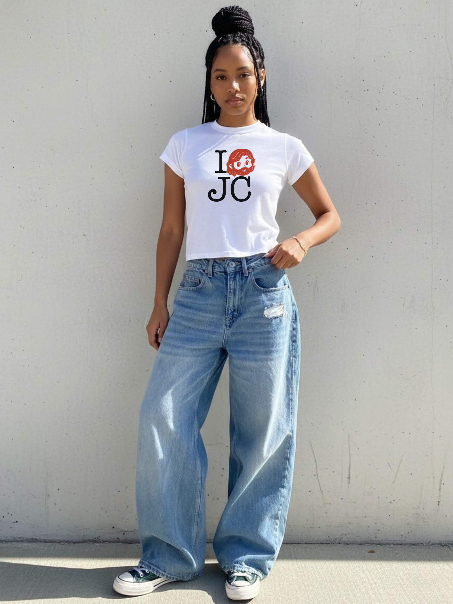 Woman wearing the SĒK Apparel I Love JC Character Baby Tee in white — faith-inspired Christian graphic tee with 'I Love JC' character design, styled with blue jeans against a gray wall