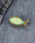 SĒK Apparel ichthys enamel pin — fish-shaped Christian lapel pin with 'JESUS' text in center, shown pinned to denim fabric