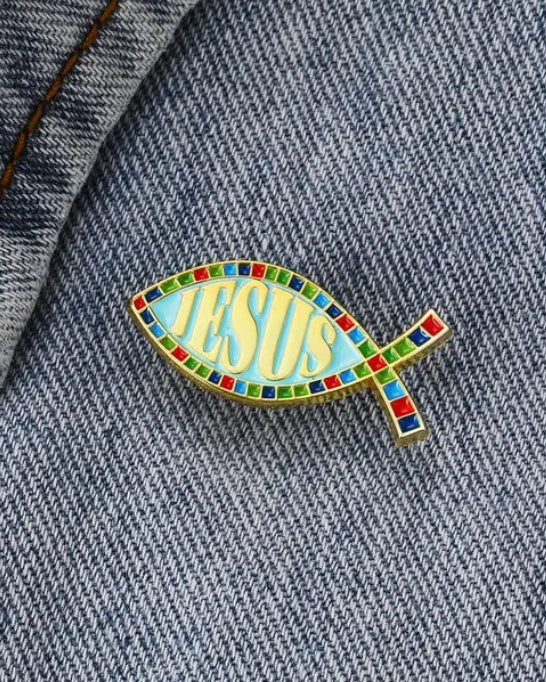 SĒK Apparel ichthys enamel pin — fish-shaped Christian lapel pin with 'JESUS' text in center, shown pinned to denim fabric