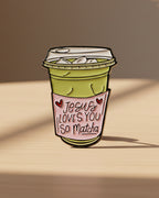 Enamel pin shaped like a matcha green tea cup with 'Jesus Loves You So Matcha' text on a beige background